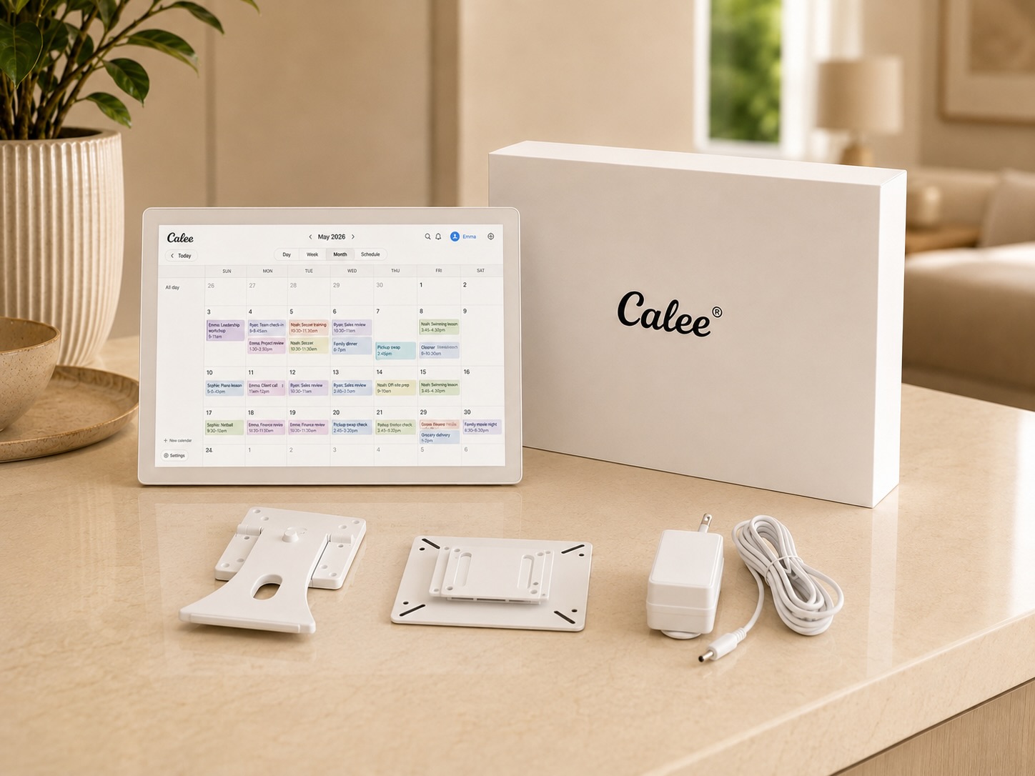 The Calee display standing on a bench showing the shared family month view, beside its Calee box, desk stand, wall-mount bracket, power adapter and cable